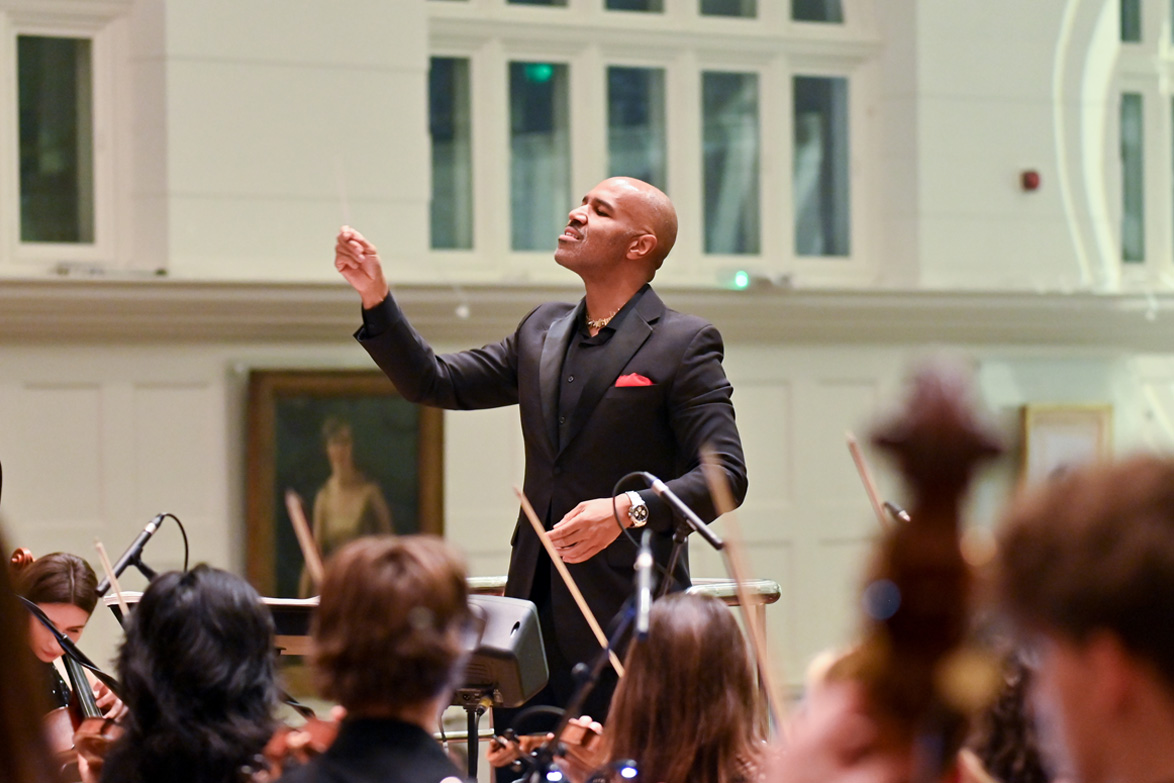 A man wearing a smart suit, leading a disco orchestra performance, with a conductors baton.